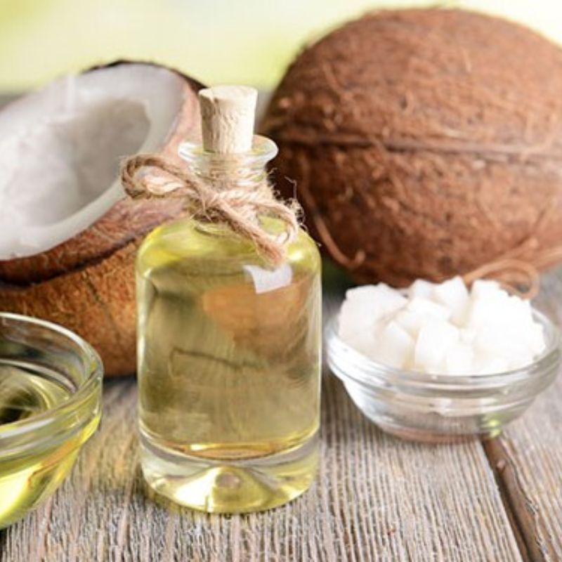A glass bottle of coconut oil with a cork, a halved coconut, and a small bowl of coconut pieces on a wooden surface.