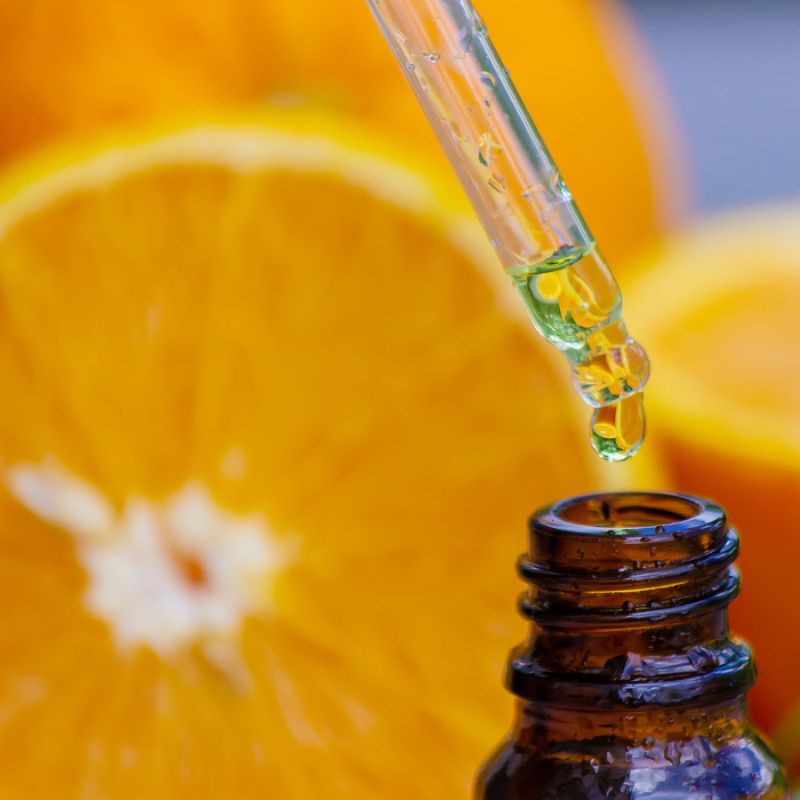 A dropper with yellow liquid above a small amber bottle, with blurred orange slices in the background.