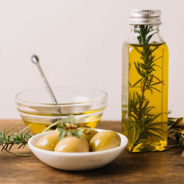 Bowl of golden olive oil, fresh olives on a plate, and a bottle of olive oil infused with rosemary.