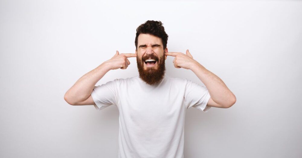 Bearded man in a white T-shirt ignoring signs by covering his ears — illustrating why men ignore yeast infection symptoms.