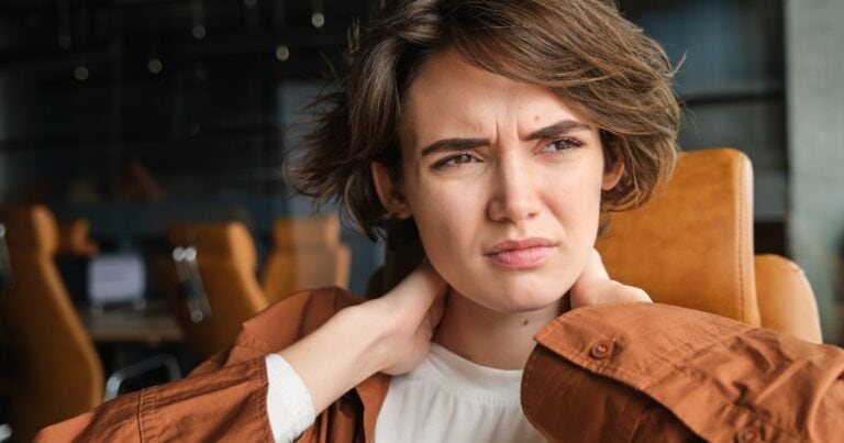 Young woman grimacing in discomfort, holding her neck, looking away from the camera — representing candida die-off (Herxheimer reaction) symptoms