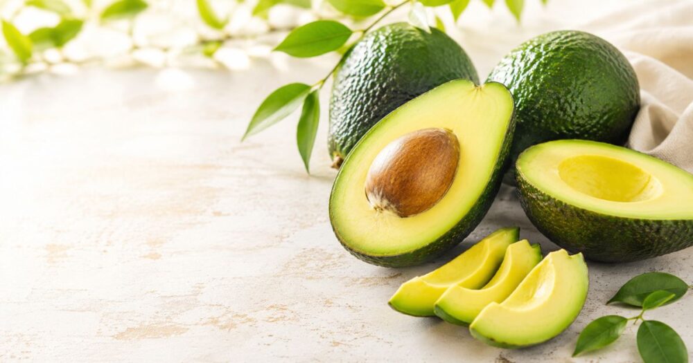 Fresh whole, halved and sliced avocados arranged on a light stone surface with green leaves and soft natural light.
