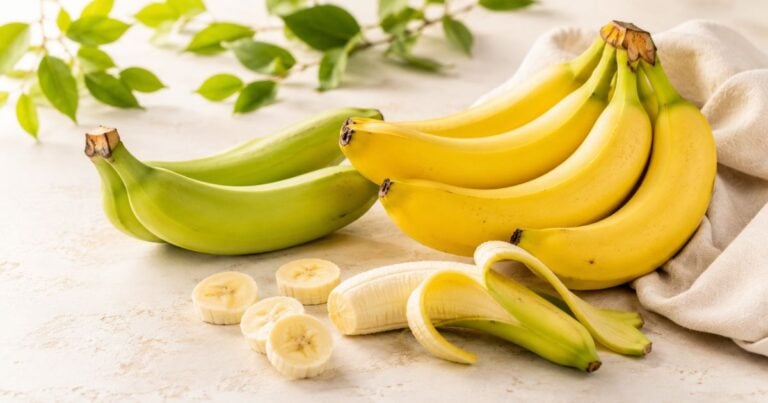 Green and ripe yellow bananas arranged on a light cream surface, with peeled banana and slices in the foreground.