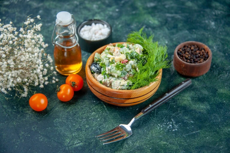A wooden bowl of antifungal candida diet vegetables and herbs, surrounded by cherry tomatoes, a bottle of oil, black pepper, a small bowl of salt, dried flowers, and a fork on a dark green surface.